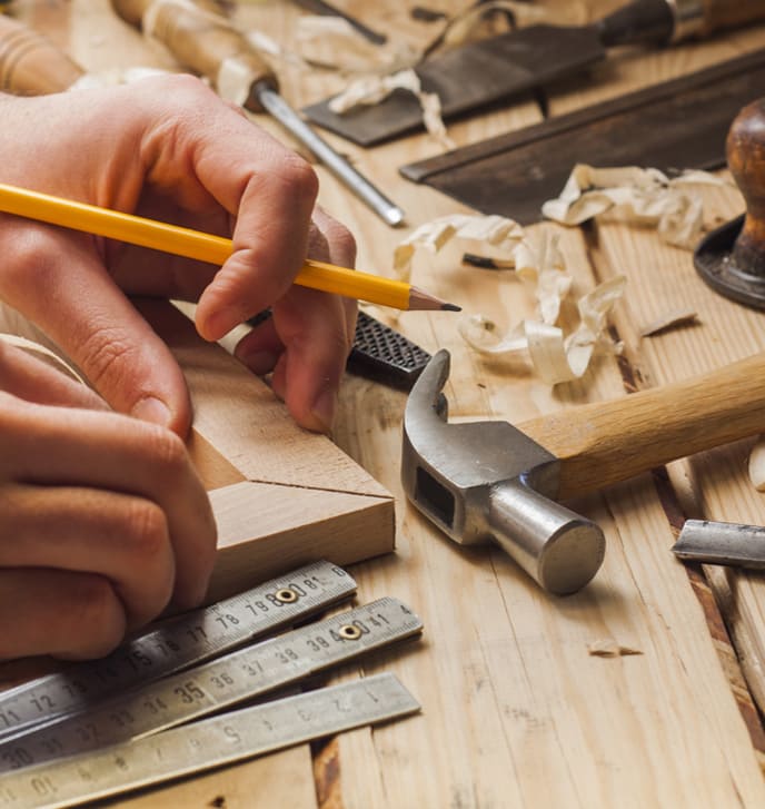 Eine Person arbeitet mit einem Bleistift und Linealen an einem Holzstück auf einem Tisch, umgeben von Werkzeugen wie Hammer, Meißel und Hobelspänen