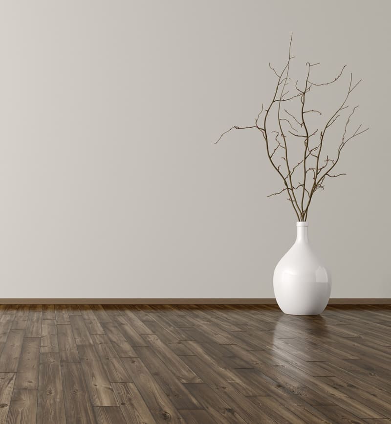 Ein minimalistischer Raum mit dunklem Holzboden und einer hellgrauen Wand, dekoriert mit einer weißen Vase, die trockene Äste enthält