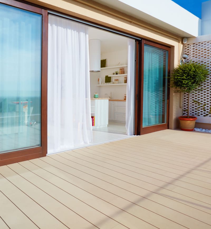 Eine helle Terrasse mit Holzfußboden, die durch eine geöffnete Schiebetür mit Glas und weißen Vorhängen mit einem modernen Innenraum verbunden ist, im Hintergrund eine Küche mit weißen Schränken und Regalen, sowie eine Topfpflanze auf der Terrasse